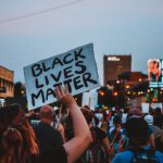 Event Changes - Back view of group of demonstrators showing banners with slogans in downtown under dark blue sky at night