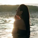 Healthy Traveling - Side view of tranquil young female tourist with long dark hair standing in waving ocean with closed eyes and enjoying summer sunset