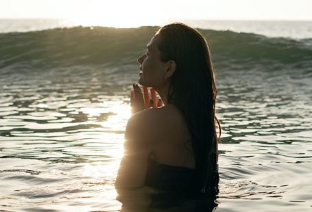 Healthy Traveling - Side view of tranquil young female tourist with long dark hair standing in waving ocean with closed eyes and enjoying summer sunset