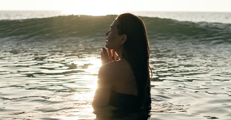 Healthy Traveling - Side view of tranquil young female tourist with long dark hair standing in waving ocean with closed eyes and enjoying summer sunset