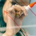 Investment Strategies - Woman in a Beige Coat Writing on a Glass Panel Using a Whiteboard Marker